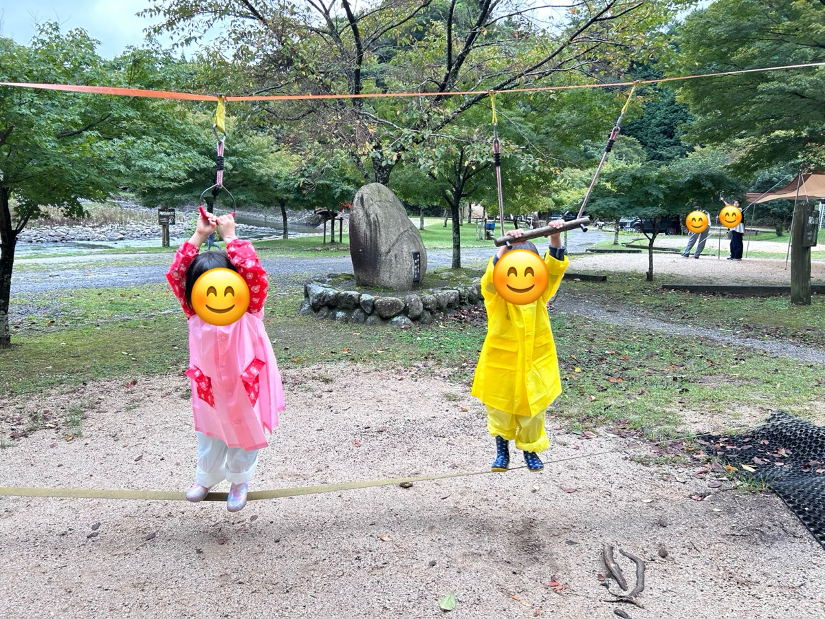 カッパ姿の子ども2人が、ロープにつかまってスラックラインに乗っているところ