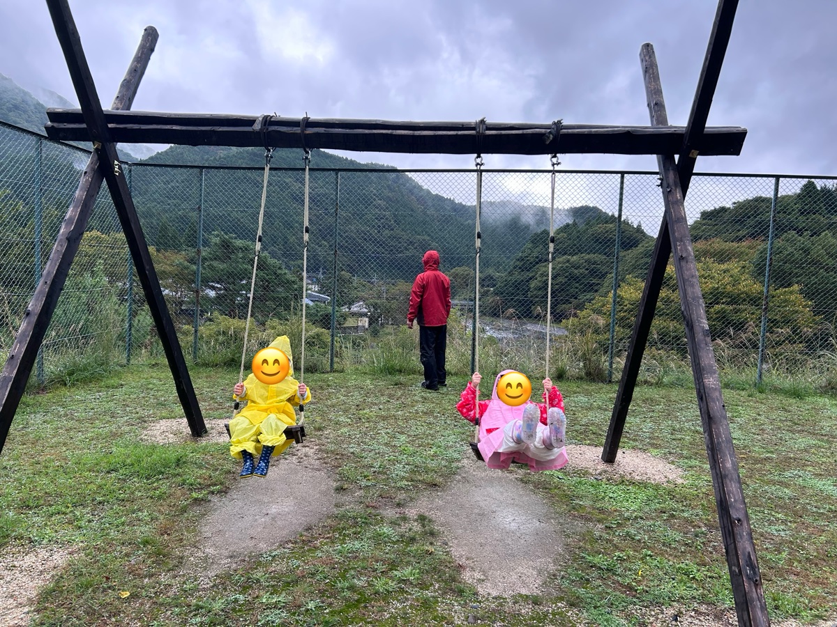 雨の日に、カッパを着た子ども2人がブランコに乗っているところ