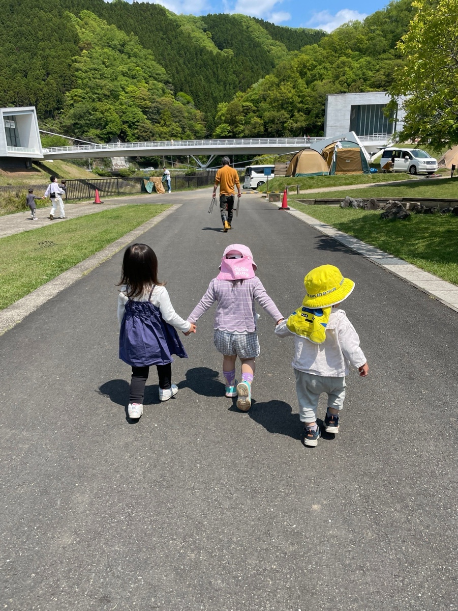 手をつないでキャンプ場の道を歩く小さな子ども3人の後ろ姿