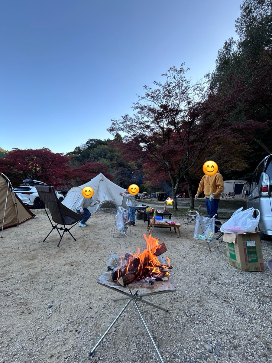 紅葉した木の下のキャンプサイト。手前に焚き火、奥にテントと家族