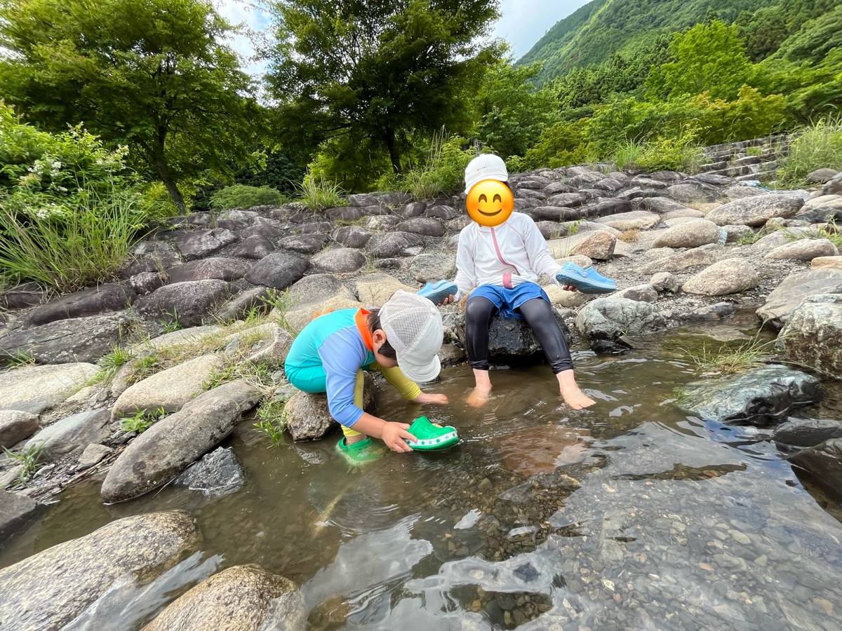川の岩場で、石に座って足を水につける子どもと、くつを洗う子ども