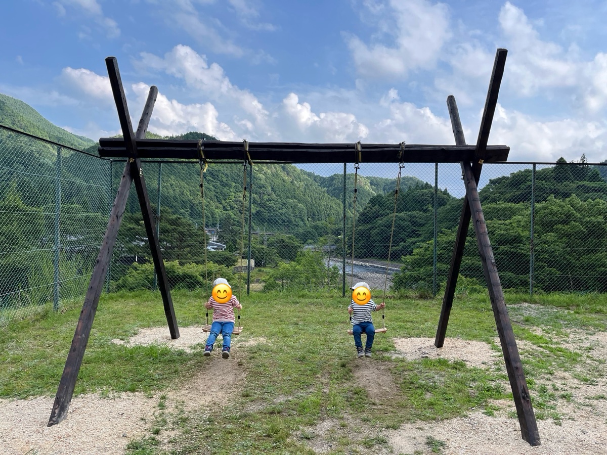 山の上に並ぶ木のブランコに乗る子ども2人。うしろに山と川が見える